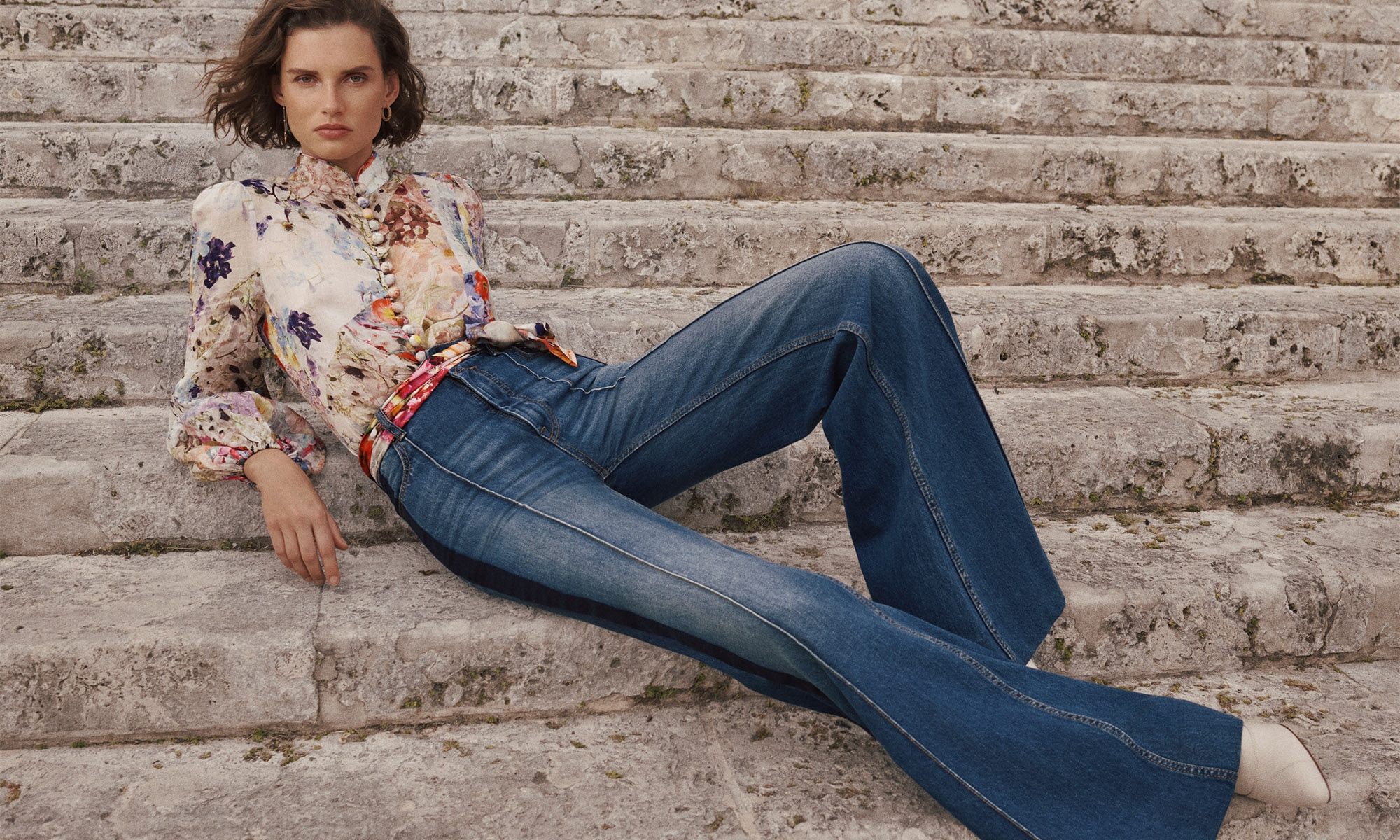 A stylish adult woman with short hair sits on stone steps wearing jeans and a floral blouse in an outdoor fashion editorial.