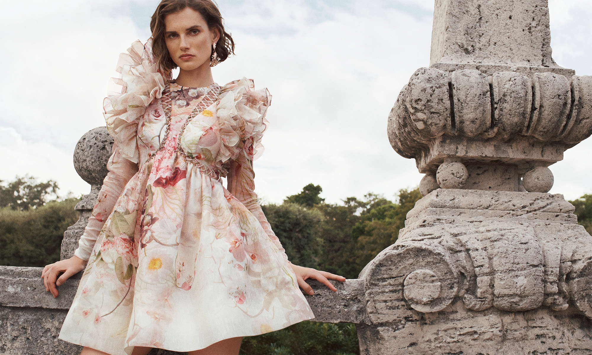 Woman in floral dress poses on a stone terrace overlooking greenery. Elegant summer fashion shoot with decorative architectural elements.