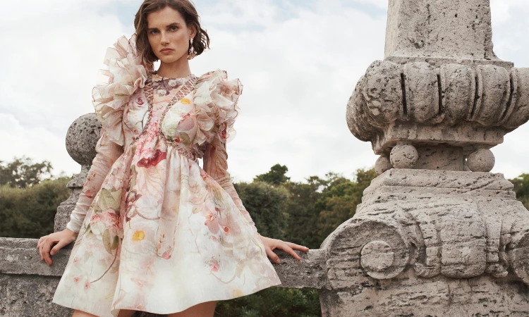 Woman in floral dress poses on a stone terrace overlooking greenery. Elegant summer fashion shoot with decorative architectural elements.