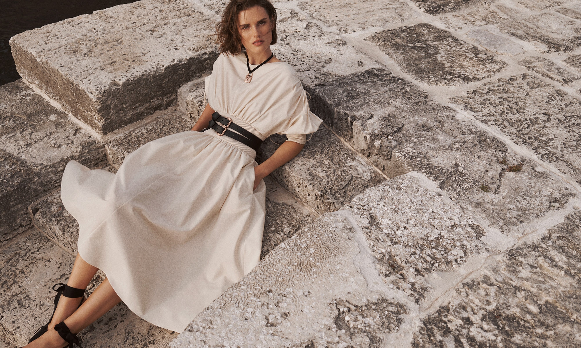 A fashion shot featuring a woman in a cream-colored dress and wide black belt, posing on stone blocks in soft lighting.