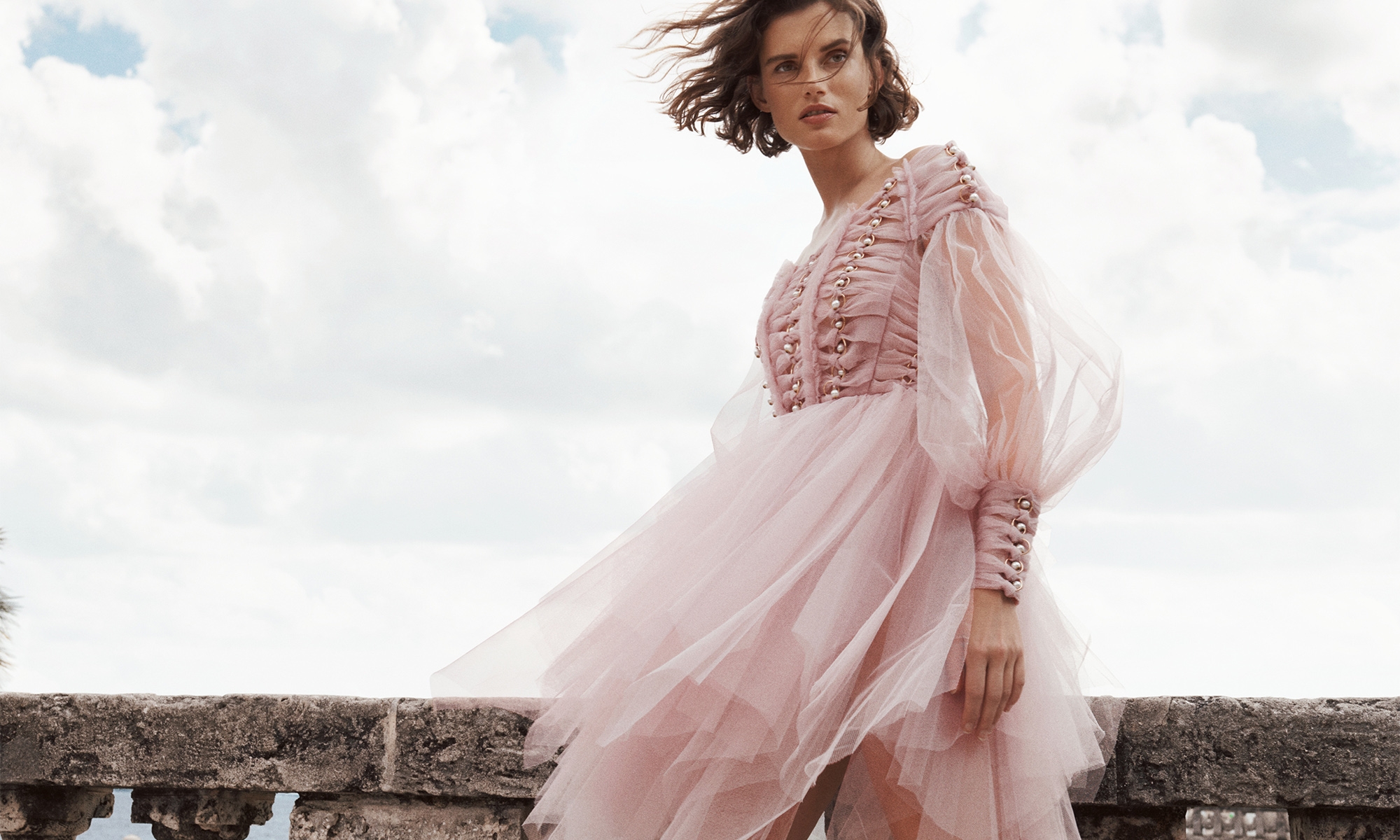 Woman in a pink tulle dress poses on a stone ledge against a sky with white clouds, creating a light and airy fashion photograph.