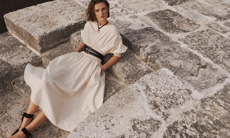Model in cream dress with black double belt seated on stone steps outdoors