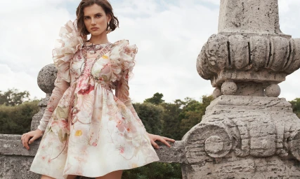 Model wearing a ruffled floral print minidress leaning against an ornate stone balustrade outdoors.