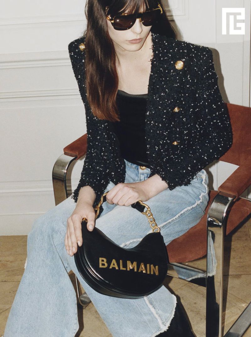 Model wearing dark sunglasses, tweed jacket, and jeans holding a black handbag while seated on a chair.