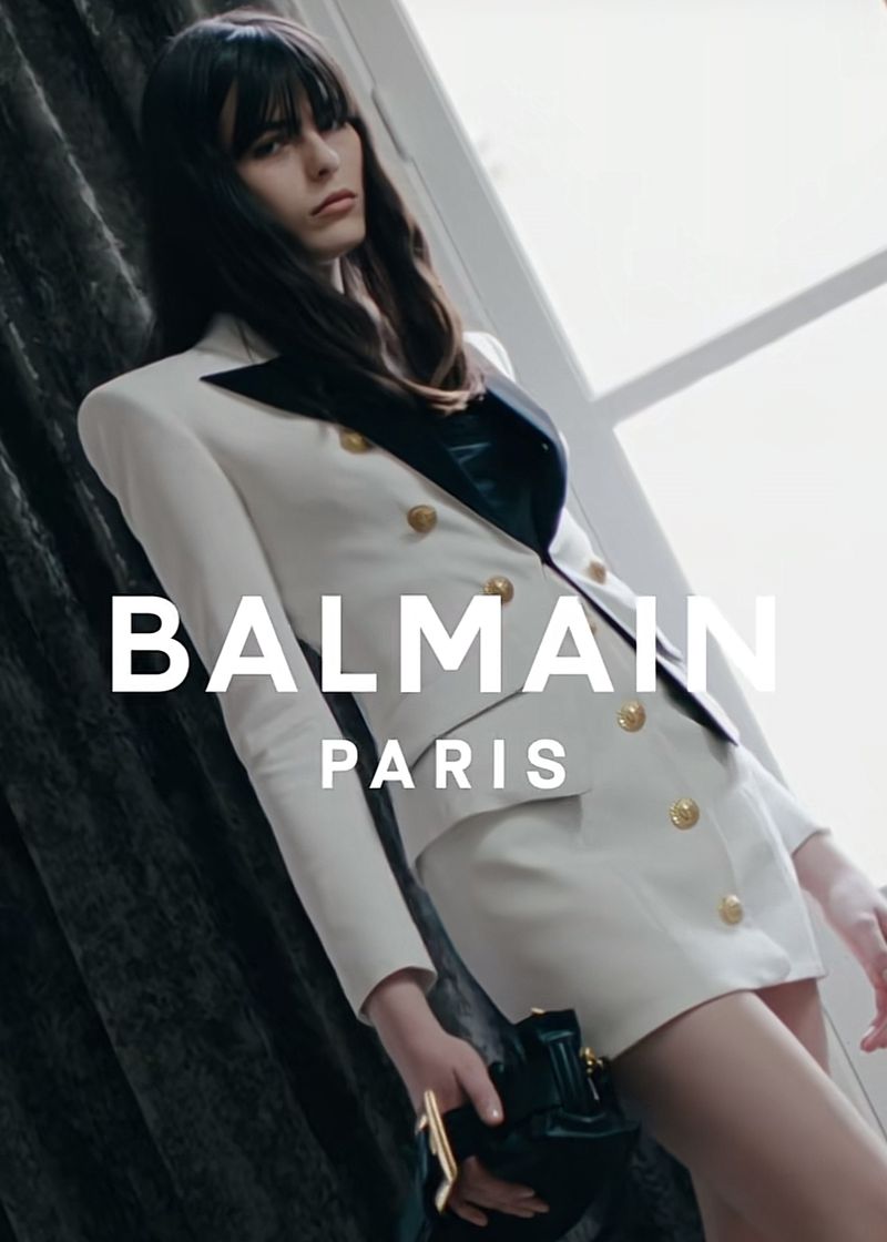 An adult female model in a white Balmain blazer dress holds a small black purse in a brightly lit indoor fashion shot.