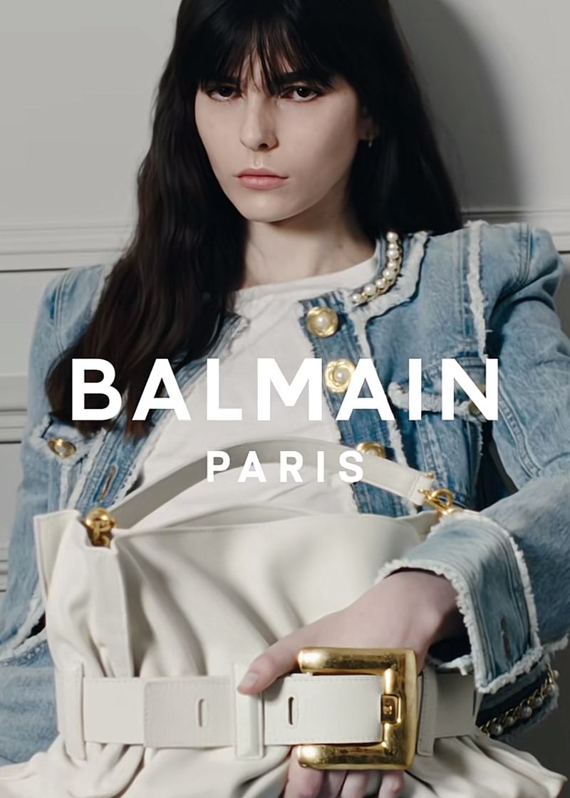 Fashion ad featuring a brunette woman in a denim jacket holding a white handbag with a gold buckle, with Balmain Paris text overlay.