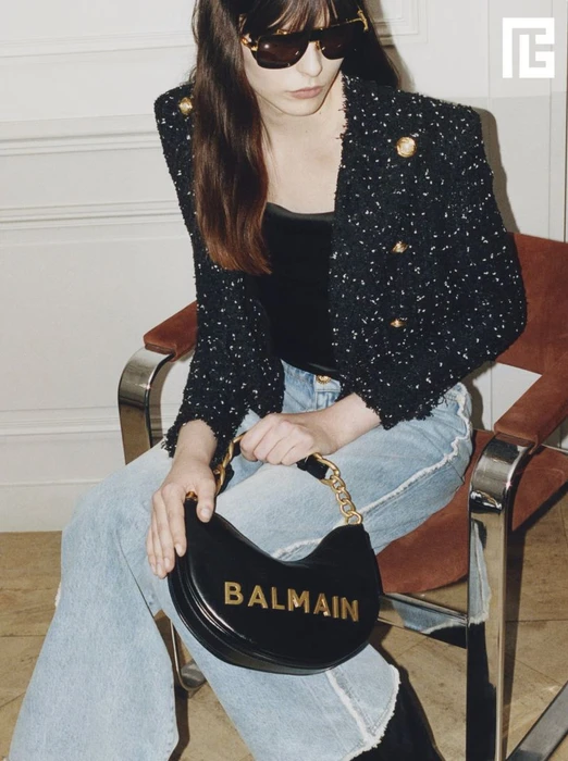 Model wearing dark sunglasses, tweed jacket, and jeans holding a black handbag while seated on a chair.