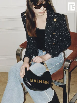 Model wearing dark sunglasses, tweed jacket, and jeans holding a black handbag while seated on a chair.
