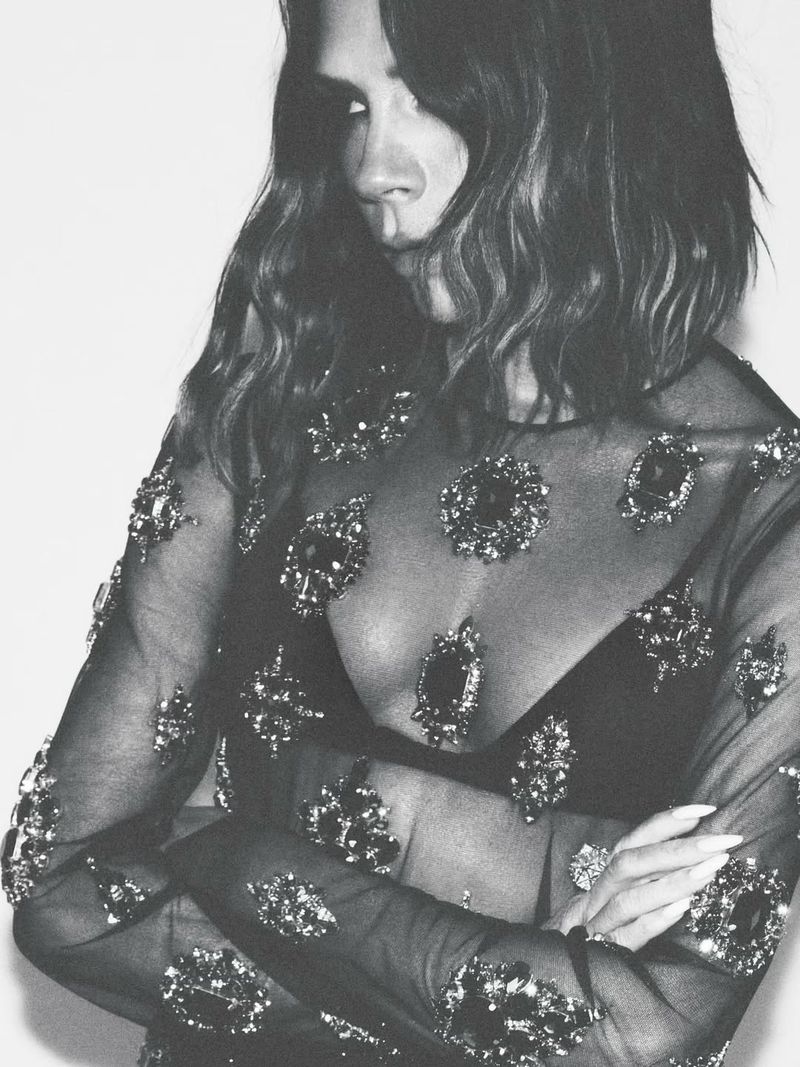 Monochrome portrait of a woman with shoulder-length wavy hair, wearing a sheer black top with bejeweled embellishments, arms crossed.