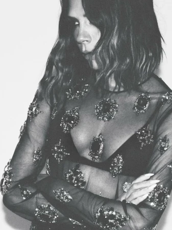 Monochrome portrait of a woman with shoulder-length wavy hair, wearing a sheer black top with bejeweled embellishments, arms crossed.