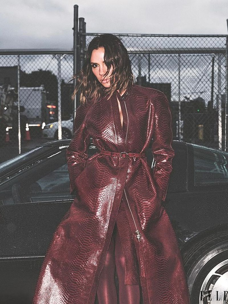 An adult woman posing in a red snakeskin trench coat next to a black car. The woman has brown shoulder-length hair.