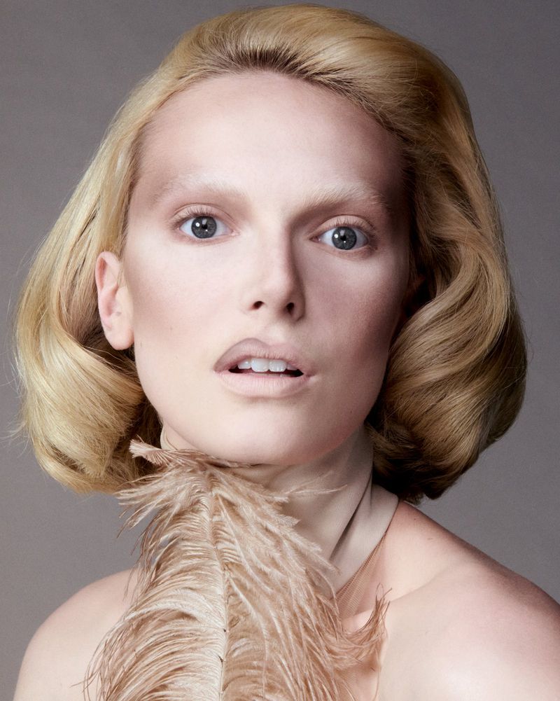 Headshot of a fair-skinned woman with styled blonde hair, light makeup, and a feather accessory, set against a neutral gray background.