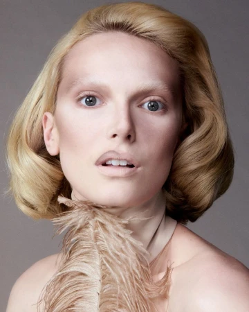 Headshot of a fair-skinned woman with styled blonde hair, light makeup, and a feather accessory, set against a neutral gray background.
