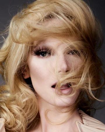 Close-up studio portrait of a blonde adult woman with long eyelashes and hair covering part of her face, against a grey background.