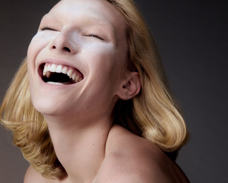 Close-up studio portrait of a smiling adult woman with blond hair and white cream covering her face, against a dark gray backdrop.