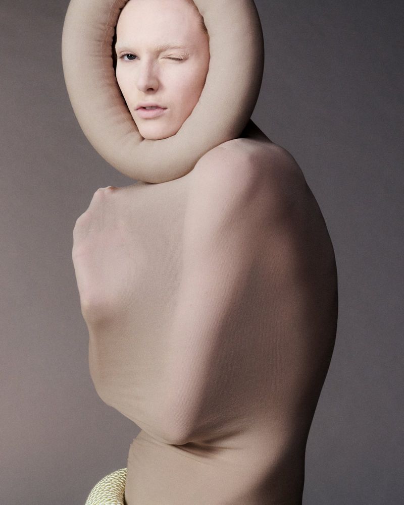 Conceptual fashion portrait of a person covered in beige fabric, winking, with circular textile collar, against a grey background.