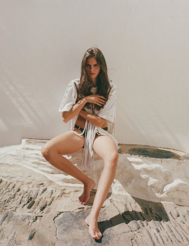 Brunette woman in an oversized t-shirt and bikini bottom sits on a stone ledge in a medium shot against a white wall backdrop.