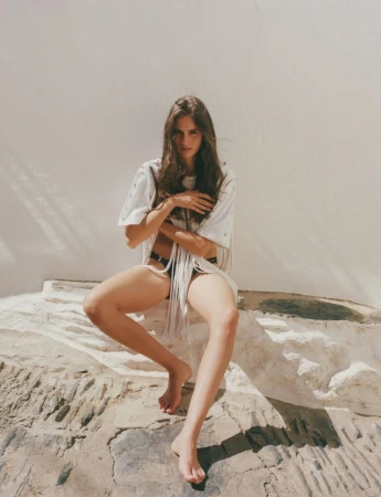 Brunette woman in an oversized t-shirt and bikini bottom sits on a stone ledge in a medium shot against a white wall backdrop.