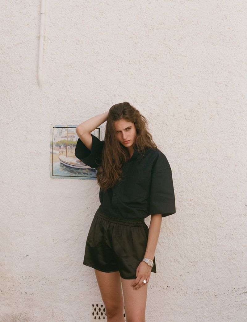 A woman with long brown hair stands against a white wall wearing a black button-down shirt and black shorts, posing and looking at the camera.