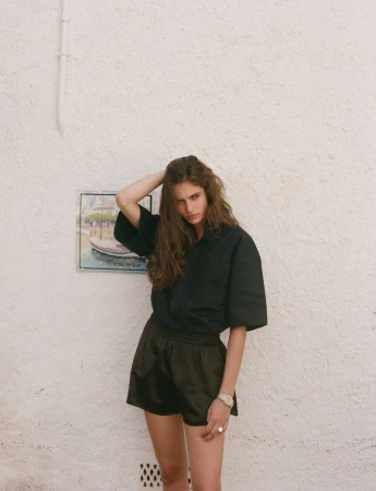 A woman with long brown hair stands against a white wall wearing a black button-down shirt and black shorts, posing and looking at the camera.
