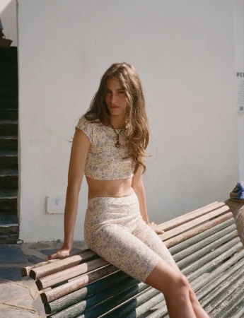 Young woman in a floral two-piece outfit sits on a weathered wooden bench against a light wall in bright daylight, fashion shoot.