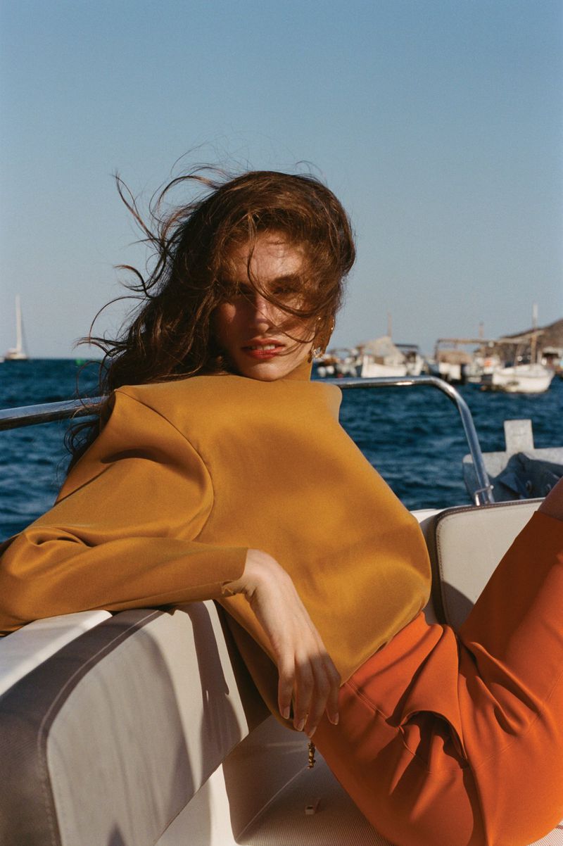 Fashion shot of a woman in an orange top and trousers sitting on a boat, with the sea and other boats in the background, shot outdoors.