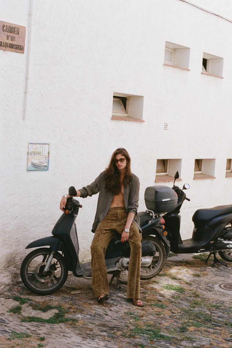 A woman with long brown hair, wearing a blazer and pants, sits on a black moped in front of a white building with windows.