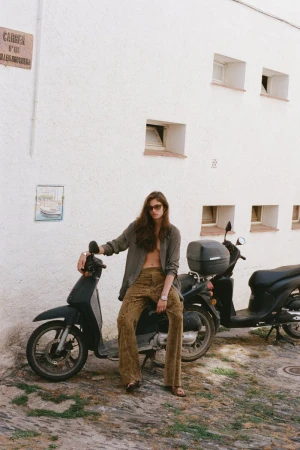 A woman with long brown hair, wearing a blazer and pants, sits on a black moped in front of a white building with windows.