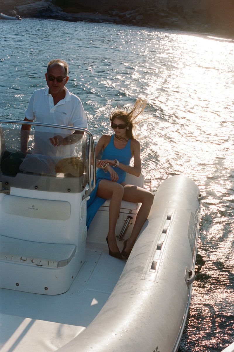 Woman in blue dress sits on the side of a boat with a man standing next to the console, in front of water and rocky cliffs.