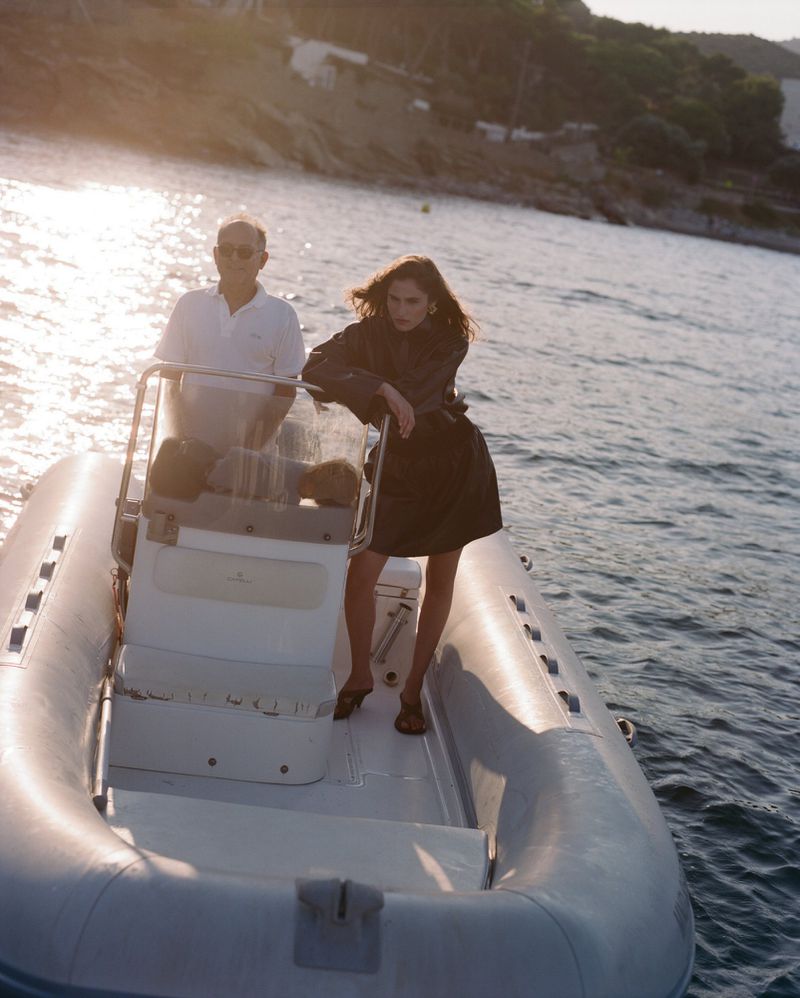 Woman in black dress on a white boat with a man driving, in sunlight on the water with landscape background. Fashion shot.