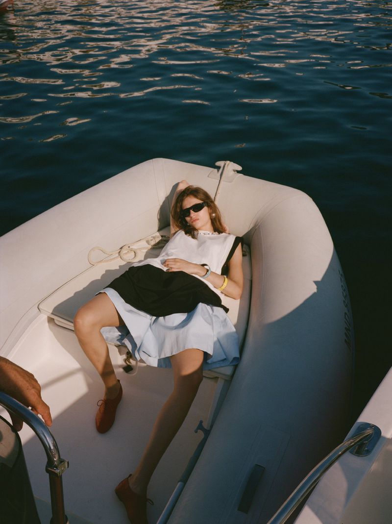 A young woman with sunglasses, wearing a white top and dark skirt, reclines on a white inflatable boat floating on dark blue water.