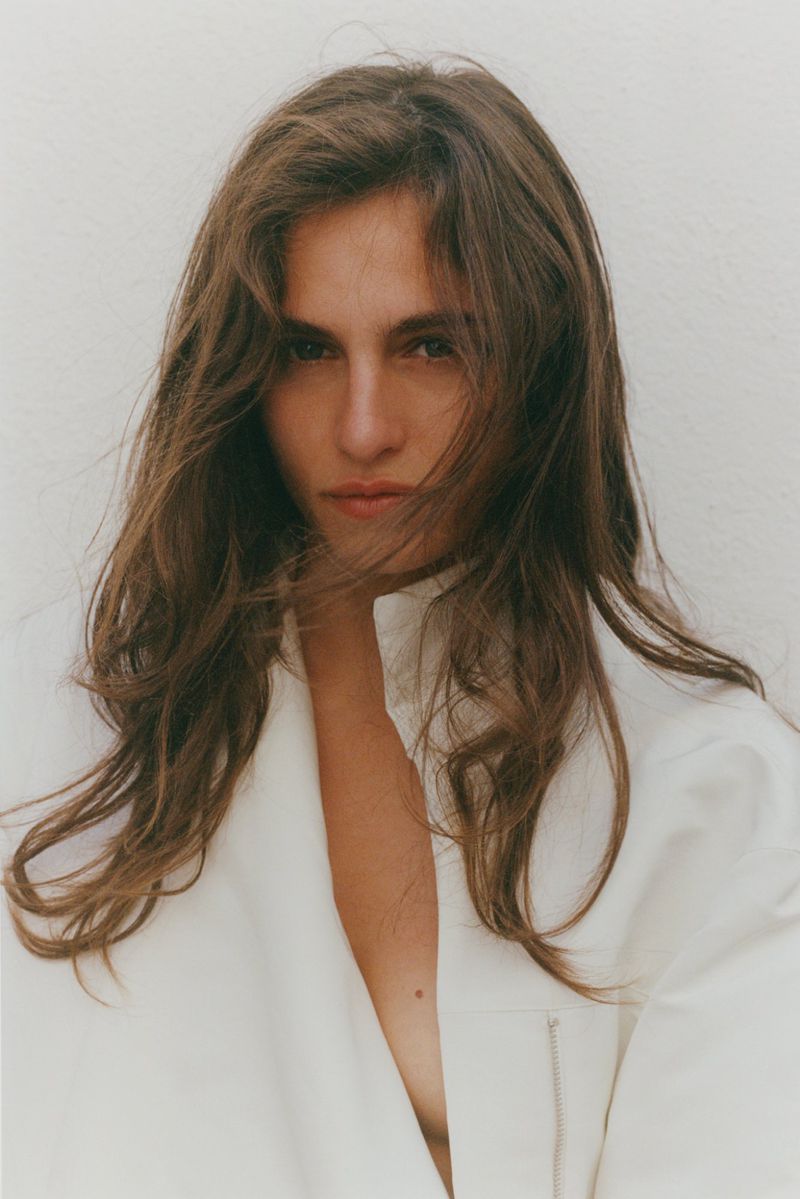 Close-up studio portrait of a woman with long brown hair, wearing a white jacket, against a white background.