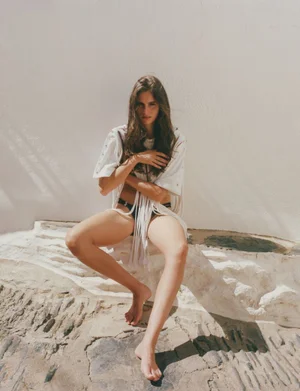 Model Nastassia Legrand seated barefoot in a white fringed top and black bikini bottoms against a sunlit whitewashed wall.