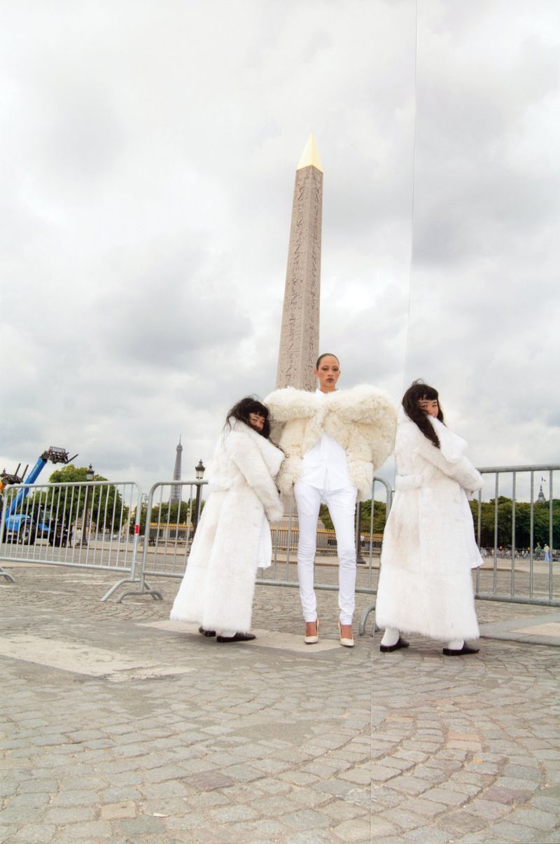 Three fashion models in white outfits pose near the Obelisk of Luxor in Paris, beneath a cloudy sky, for a fashion editorial.
