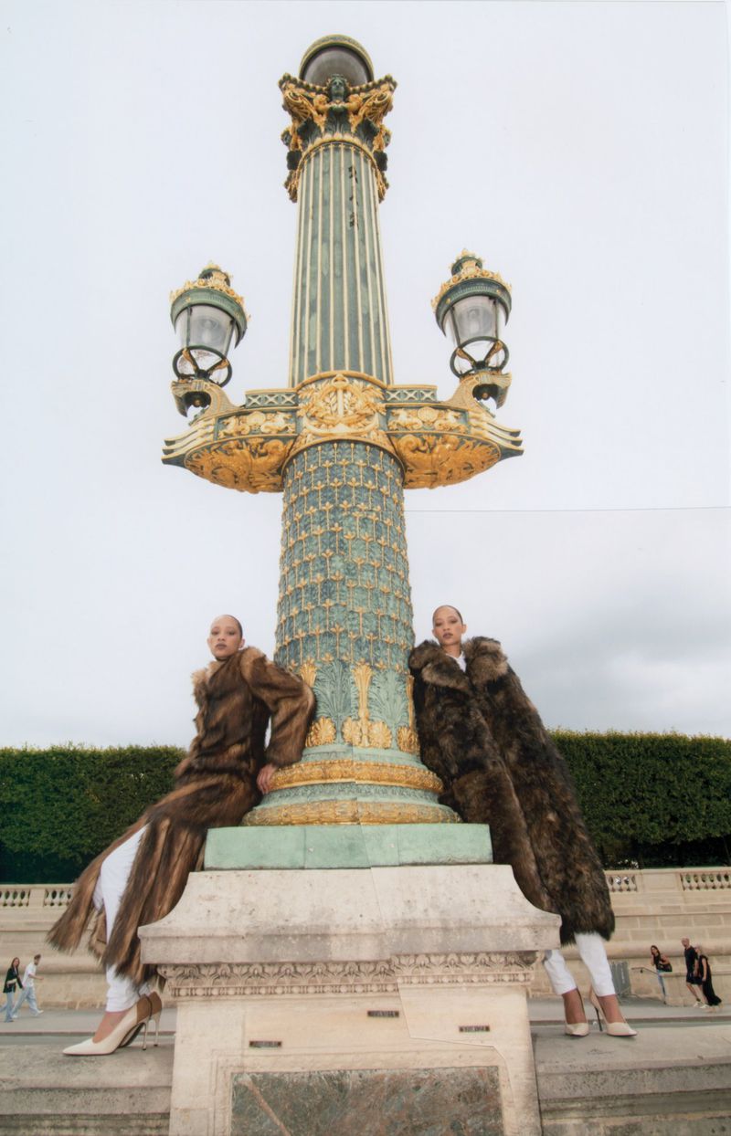 Two women in fur coats pose by a vintage lamp post in an urban setting. The scene is captured with a low-angle shot.