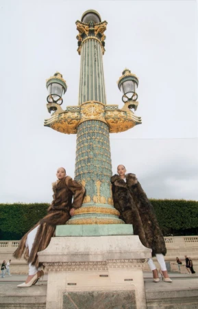Two women in fur coats pose by a vintage lamp post in an urban setting. The scene is captured with a low-angle shot.