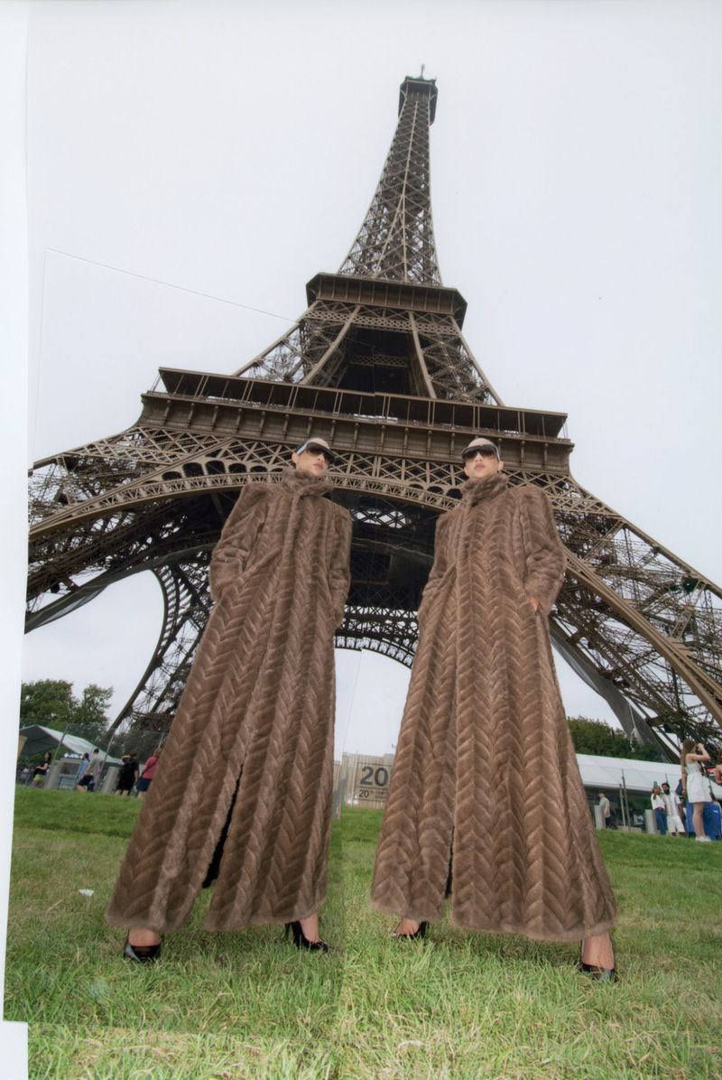 Two women in long brown coats pose in front of the Eiffel Tower on a cloudy day, showcasing fashion and travel in Paris.