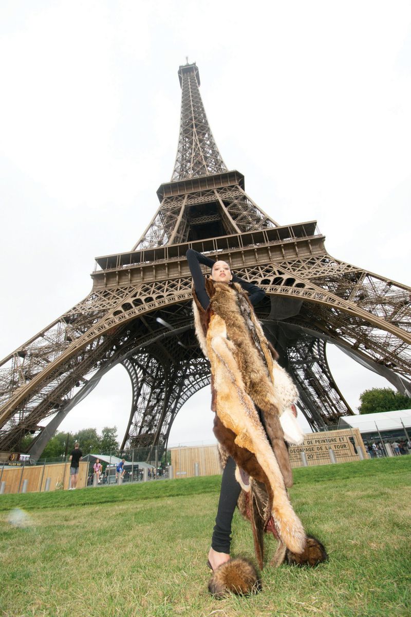 Woman wearing a fur coat poses in front of the Eiffel Tower in Paris, France on a grassy lawn under an overcast sky.