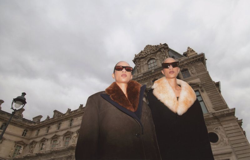 Two women in oversized coats and sunglasses stand in front of an ornate building under a cloudy sky, emphasizing fashion and architecture.
