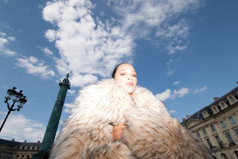 Woman in a fur coat stands in an urban square with a column, historic buildings, and a blue sky with clouds in the background.