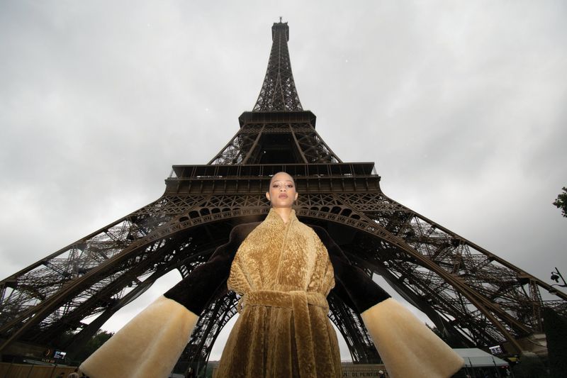 Woman in a plush coat poses in front of the Eiffel Tower on a cloudy day. Tourism and fashion photography in Paris, France.
