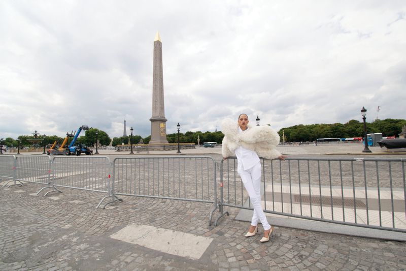Woman in white outfit and feather boa poses in Paris with the Luxor Obelisk and metal barriers on a cloudy day.