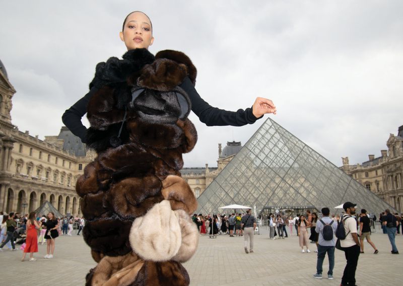 Adult female poses in fur coat at Louvre courtyard, Paris, with pyramid, architecture and crowd of tourists in the background.