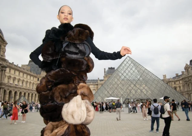 Adult female poses in fur coat at Louvre courtyard, Paris, with pyramid, architecture and crowd of tourists in the background.