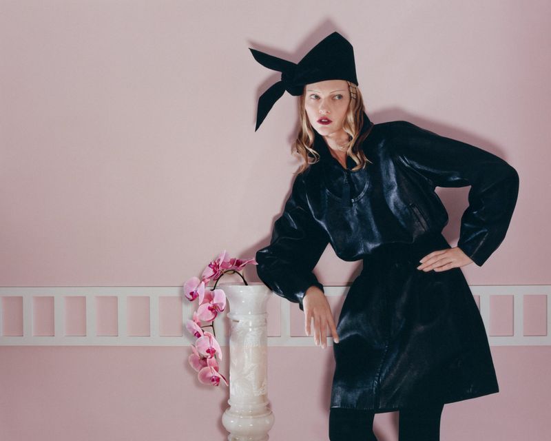 A fashionable adult female model poses indoors, wearing a black hat and black leather-like dress next to a white column with pink flowers.