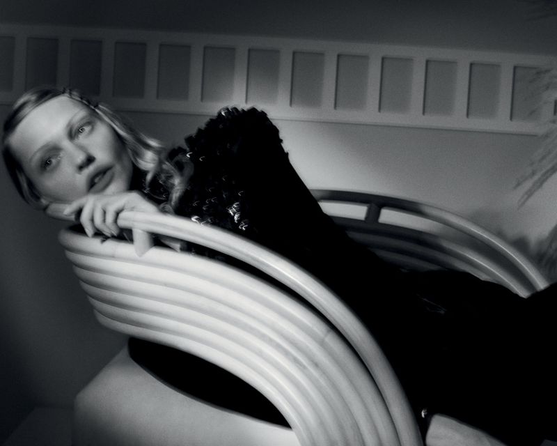 Black and white portrait of a blond woman reclining on a modern white lounge chair in an indoor setting with geometric background.