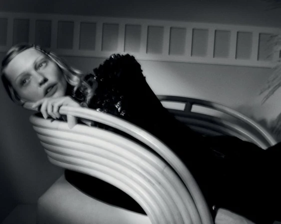 Black and white portrait of a blond woman reclining on a modern white lounge chair in an indoor setting with geometric background.