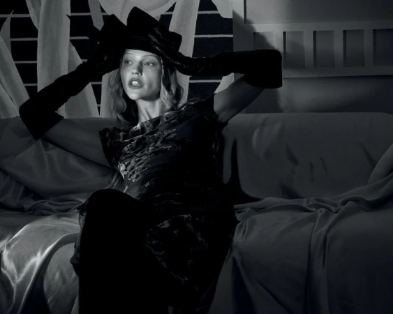 Black and white portrait of a woman wearing a hat and gloves, seated on a couch with dramatic lighting and soft textures.
