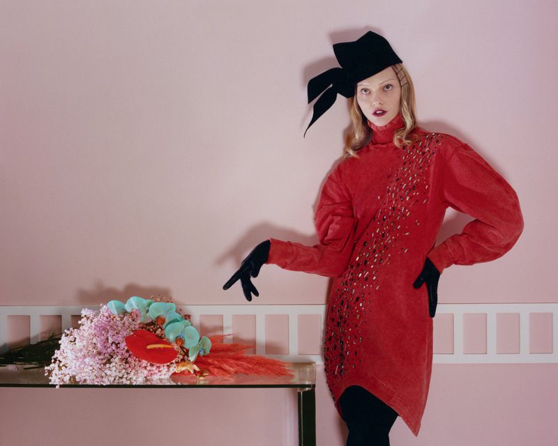 A model poses in a red, embellished dress and black hat next to flowers on a glass table against a pink wall in a studio setting.