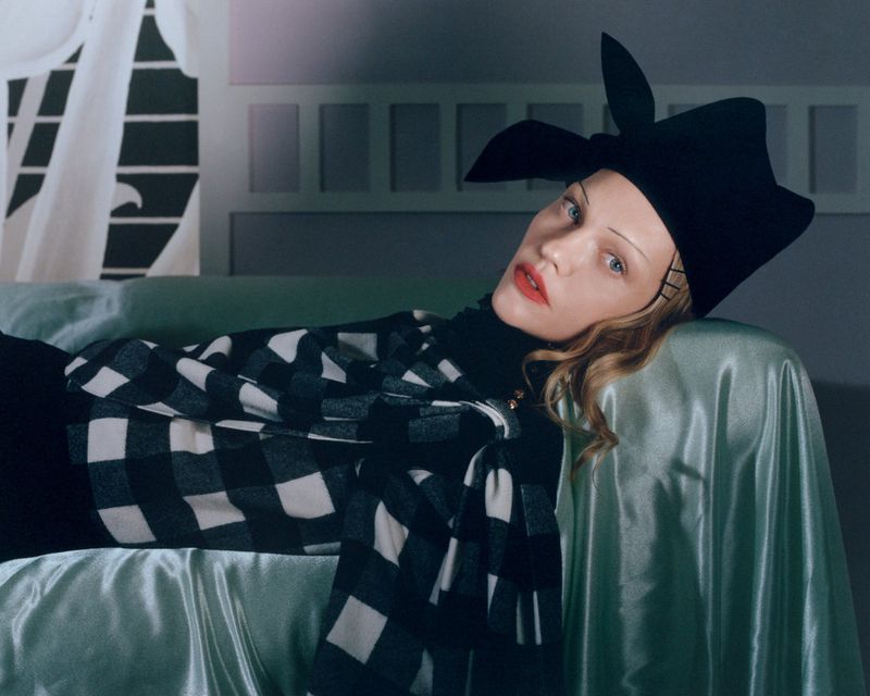 Adult female with curly blonde hair lounges with a black and white buffalo check shirt and black hat on a satin-covered surface.